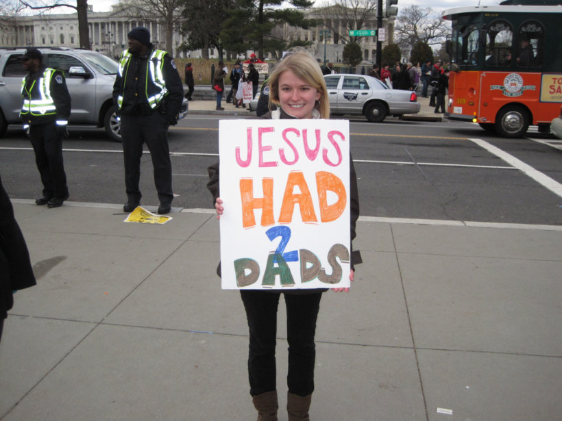 woman holding Jesus Had 2 Dads sign on sidewalk