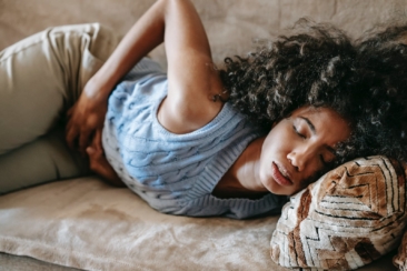 High angle of African American woman with afro hairstyle and closed eyes having pain in belly lying on sofa
